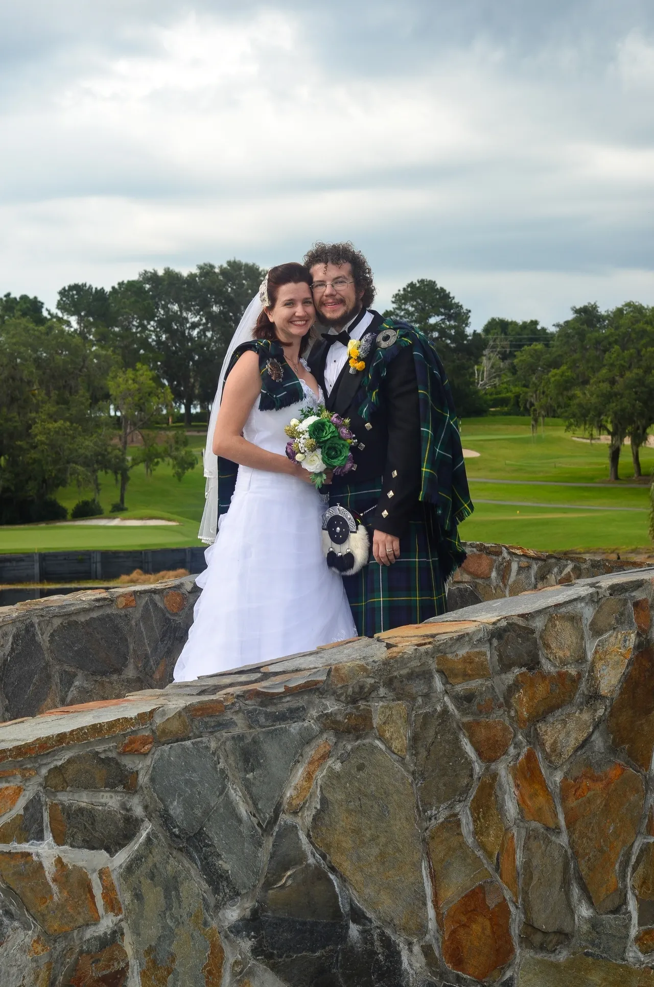 A couple in wedding attire, including a traditional kilt, posing on a stone bridge overlooking a golf course.