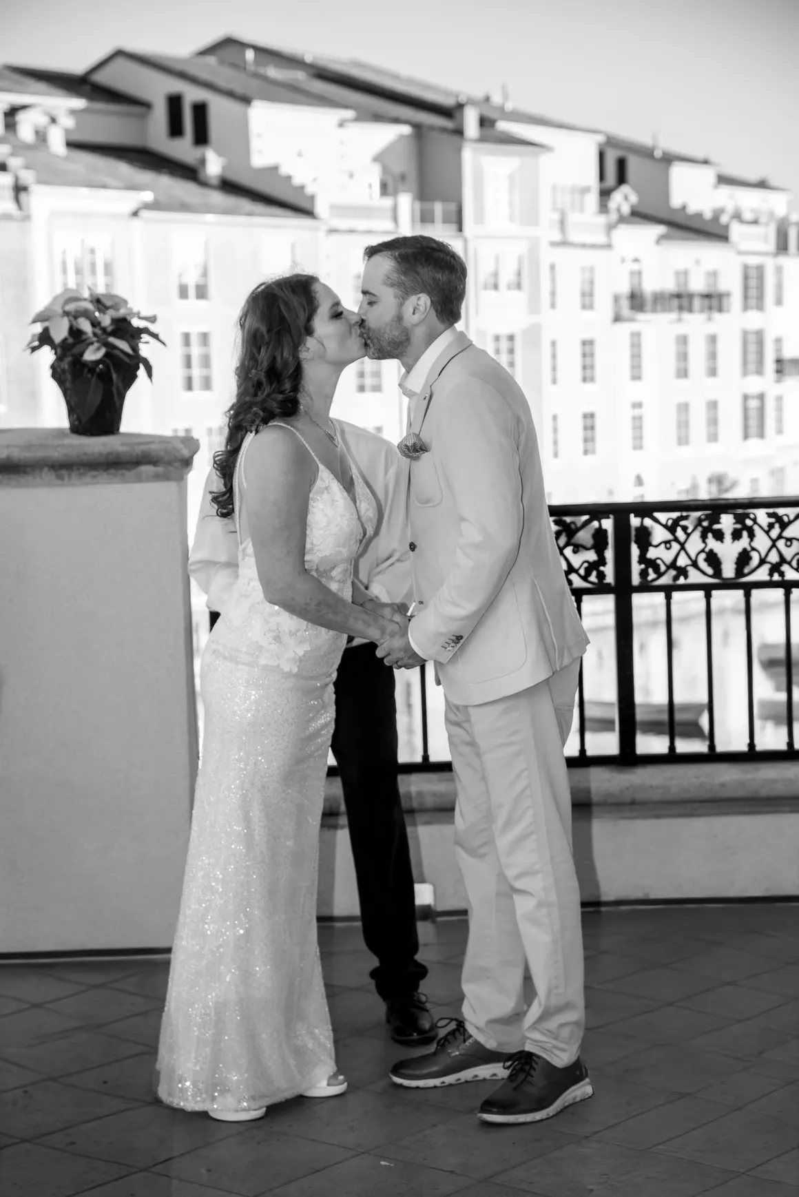 A newlywed couple kisses on a balcony at sunset, with an officiant standing between them against a building background.