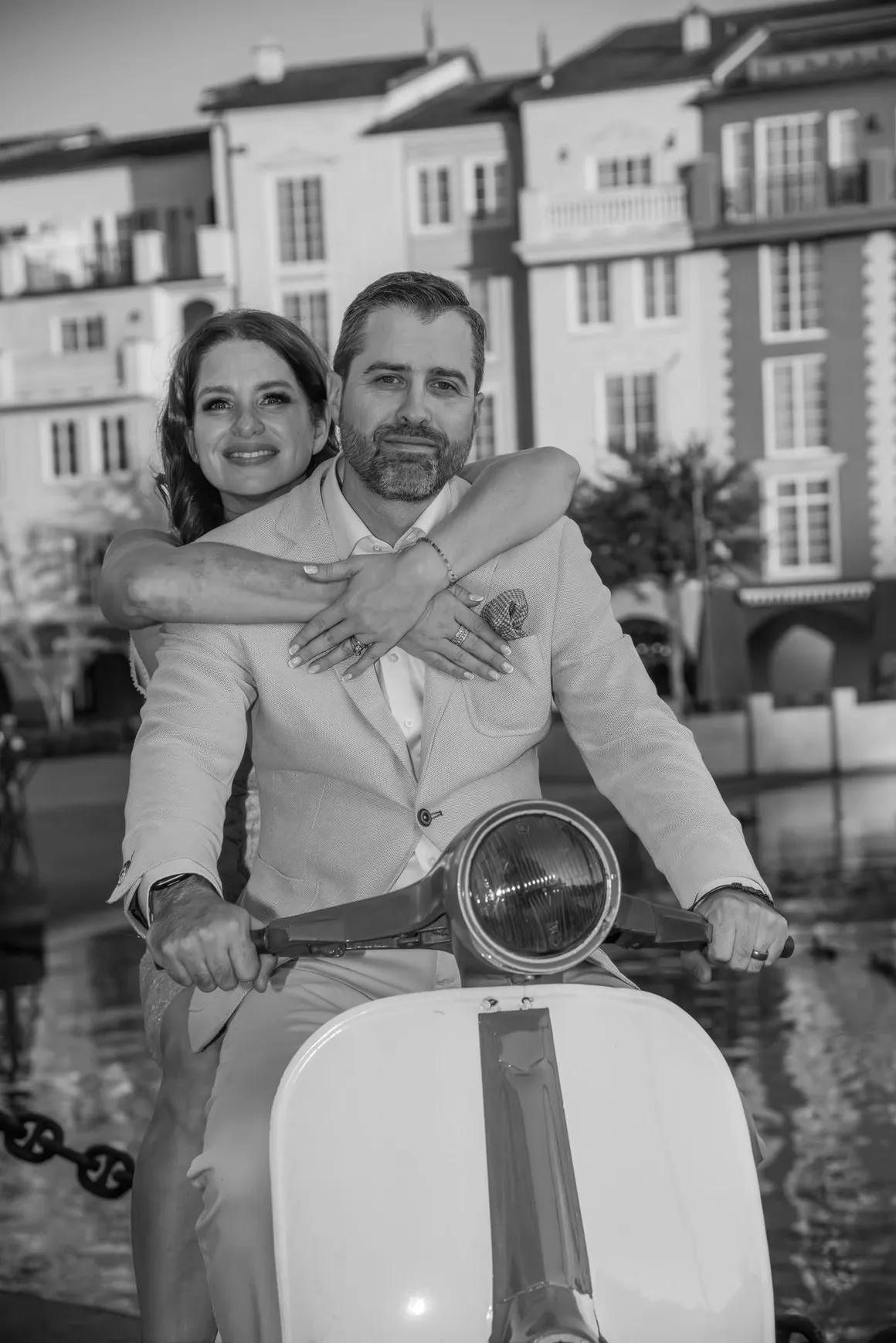 A black-and-white photo of two people hugging on a scooter in front of European-style buildings near a canal.
