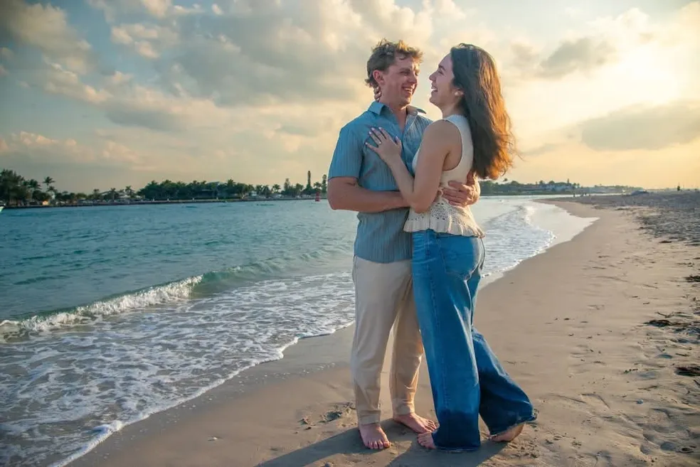 A couple stands close, laughing and holding each other on a beach at sunset by the water’s edge.