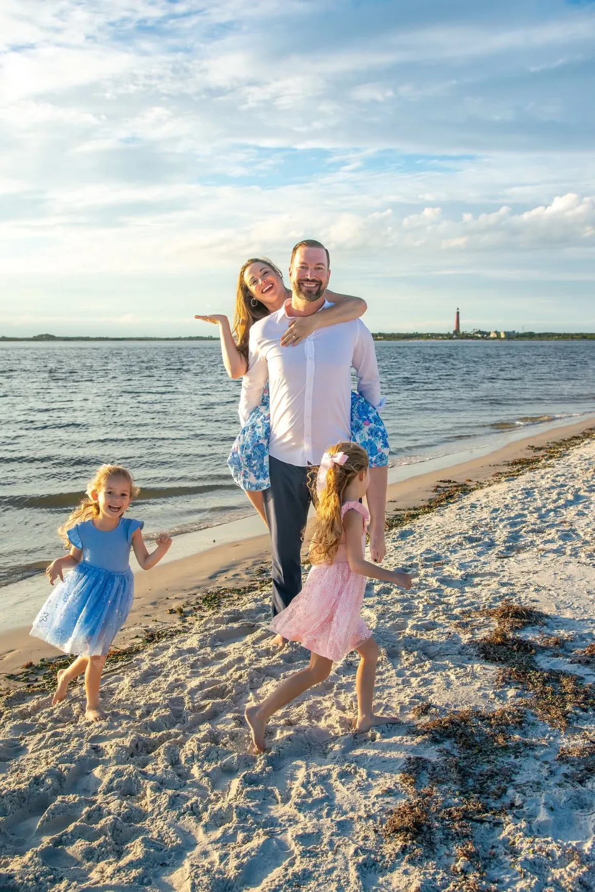 A family of four on a beach at sunset, with parents embracing and two children running on the sand.