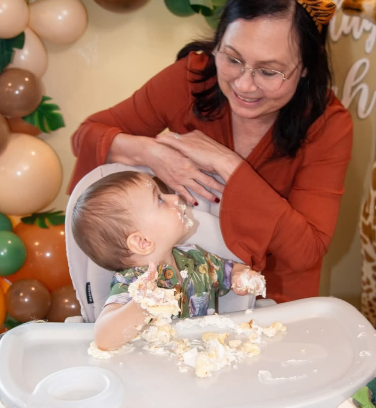 An adult in an orange shirt watches a toddler with cake on their face and hands sitting at a high chair by balloon decor.