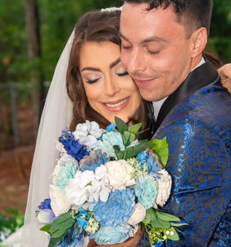 A bride and groom in a floral-patterned blue suit embrace, holding a large bouquet of blue and white flowers.