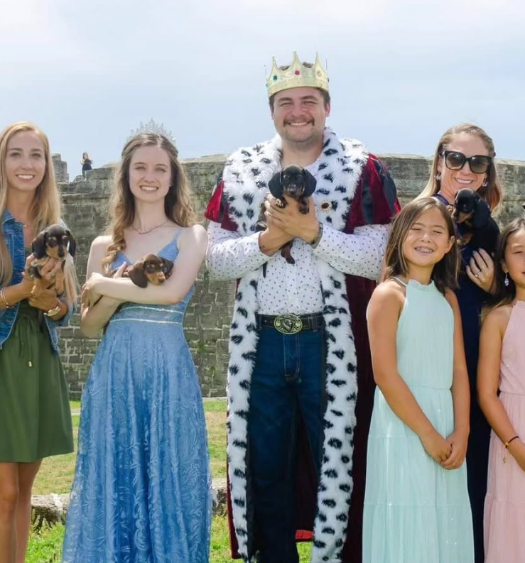 A group posing outdoors, including a person in a crown and velvet robe, each holding a small dark puppy.