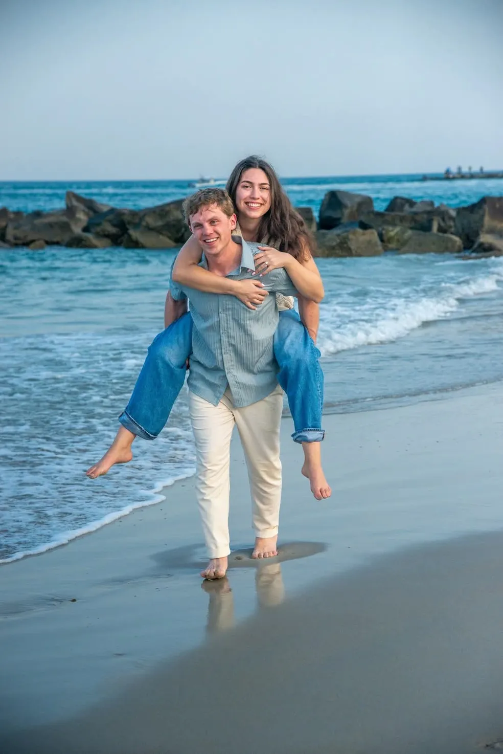 A smiling person gives another person a piggyback ride along the shoreline, with rocks and ocean in the background.