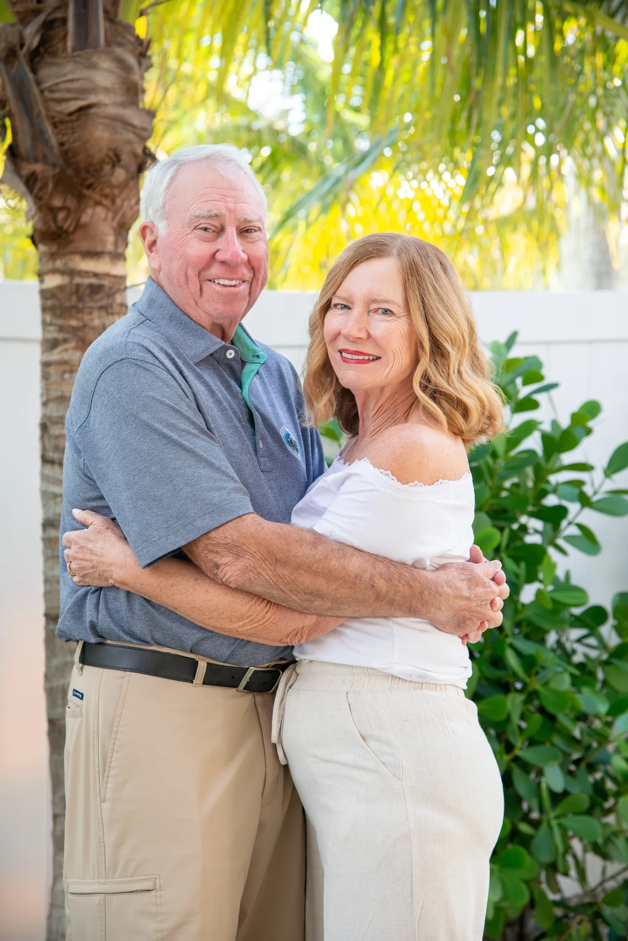 A smiling couple embraces outdoors, standing in front of a palm tree and green foliage against a white wall.