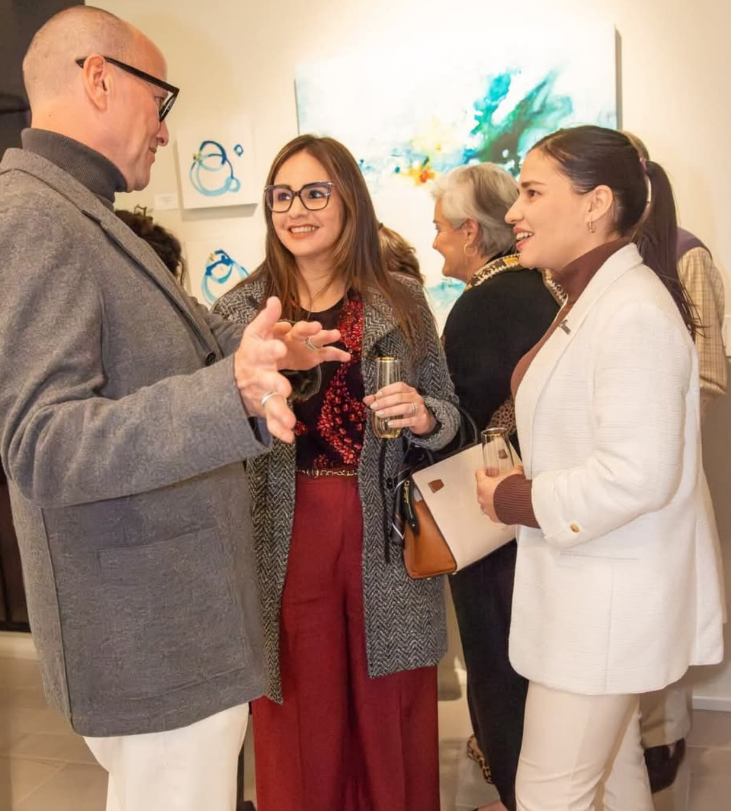 Three people in an art gallery discuss abstract paintings; one person gestures while speaking to the others holding drinks.