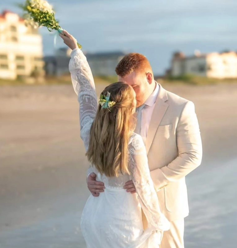 A person in a white wedding dress holds a bouquet aloft while embracing a person in a tan suit on a sunny beach.