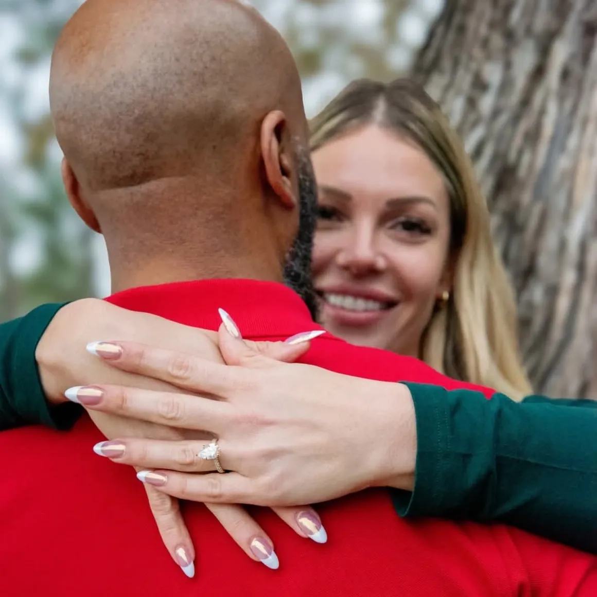 A woman with a ring on her finger embraces a person from behind, with both smiling in an outdoor setting.