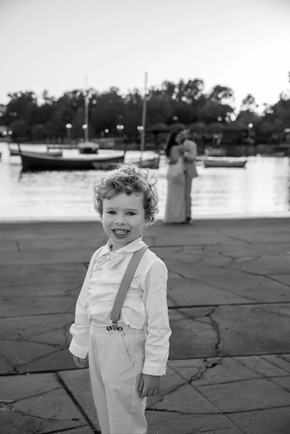 A smiling child in a light-colored outfit with suspenders stands in the foreground, with an out-of-focus couple by water.