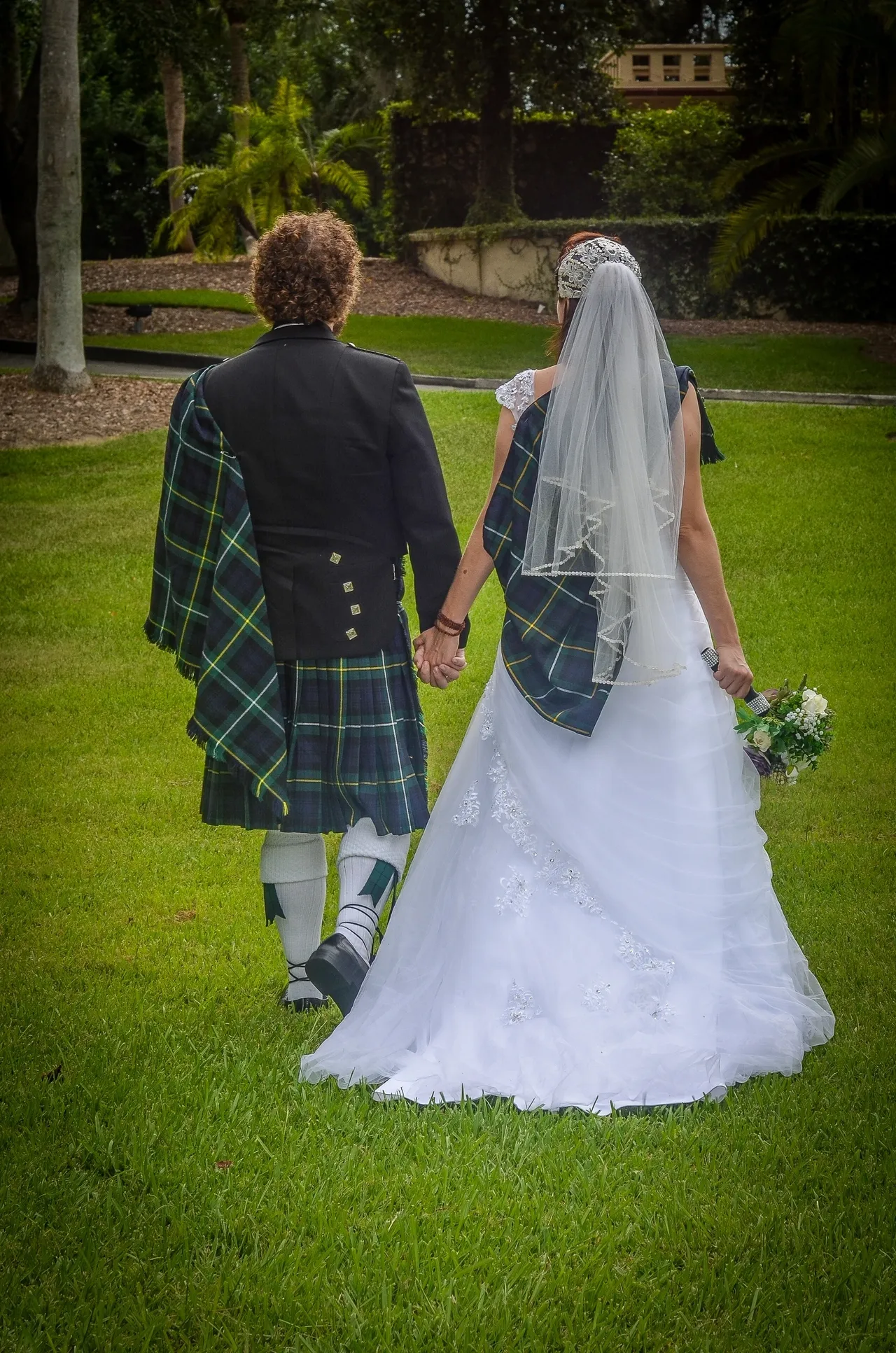 A couple in traditional Scottish wedding attire, wearing matching tartan sashes, walking hand-in-hand across a grassy lawn.