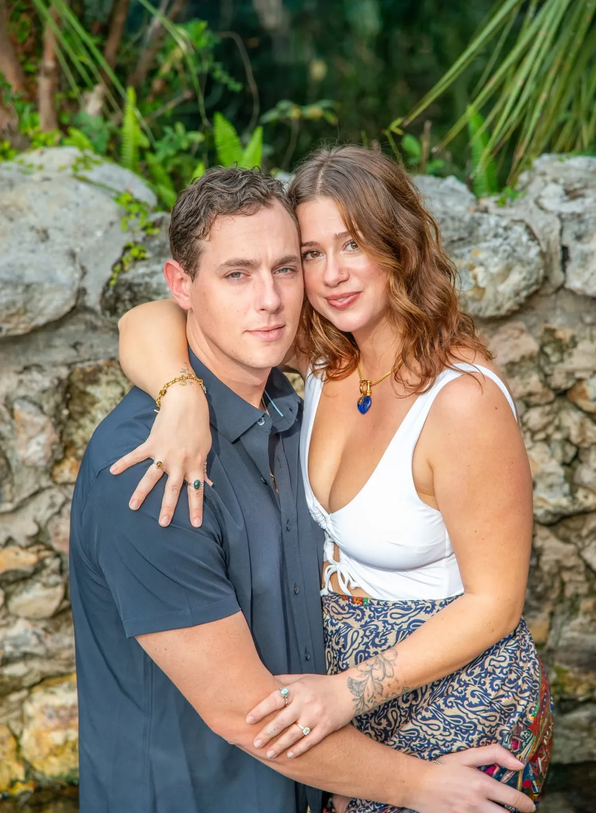 A couple embraces closely in front of a stone wall, smiling softly toward the camera.