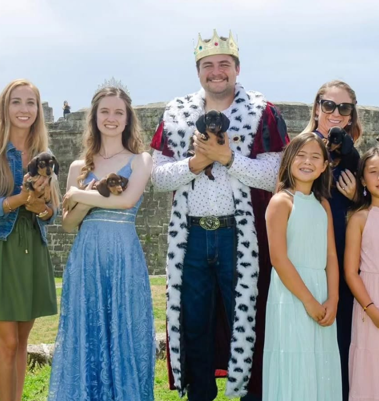 A group of people, including one wearing a crown and robe, pose together outdoors, some holding small puppies.