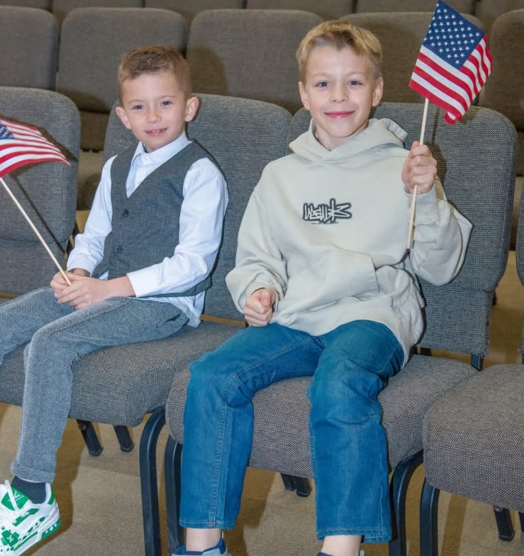 Two children sit in chairs in a room, both smiling while holding small American flags.