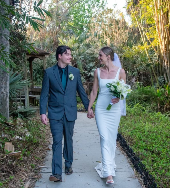 A couple in wedding attire walks hand-in-hand along a path in a garden, smiling at each other.