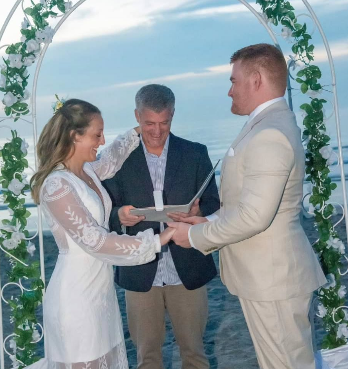 A couple holds hands during a beach wedding ceremony, with an officiant standing between them under a floral arch.