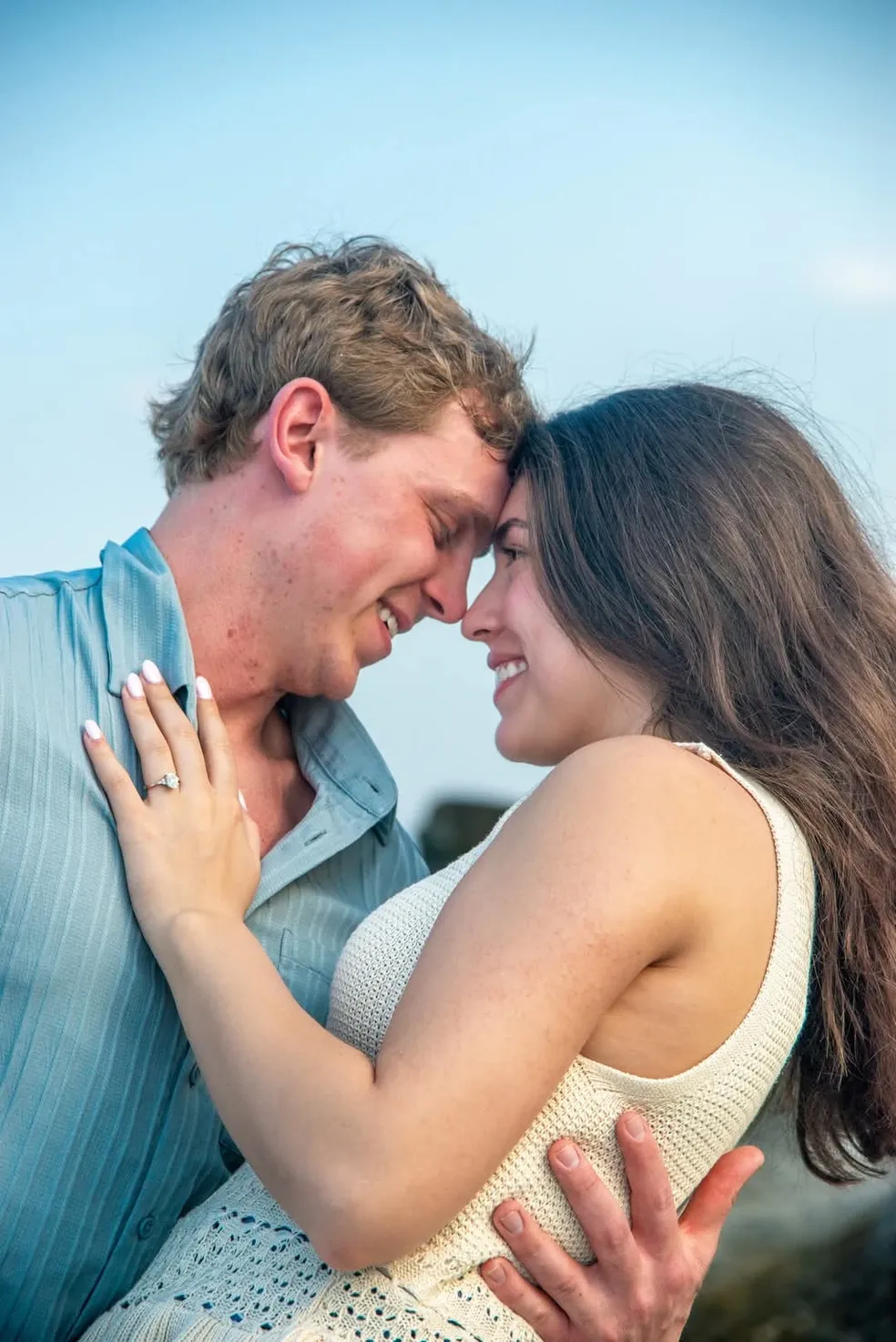 A couple gazes lovingly at each other, holding one another against a clear, light blue sky.