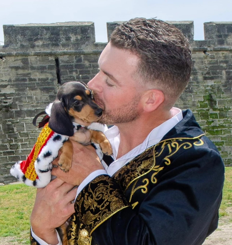 A person in a decorative black and gold jacket kisses a dachshund puppy wearing a small royal cape in front of a stone wall.