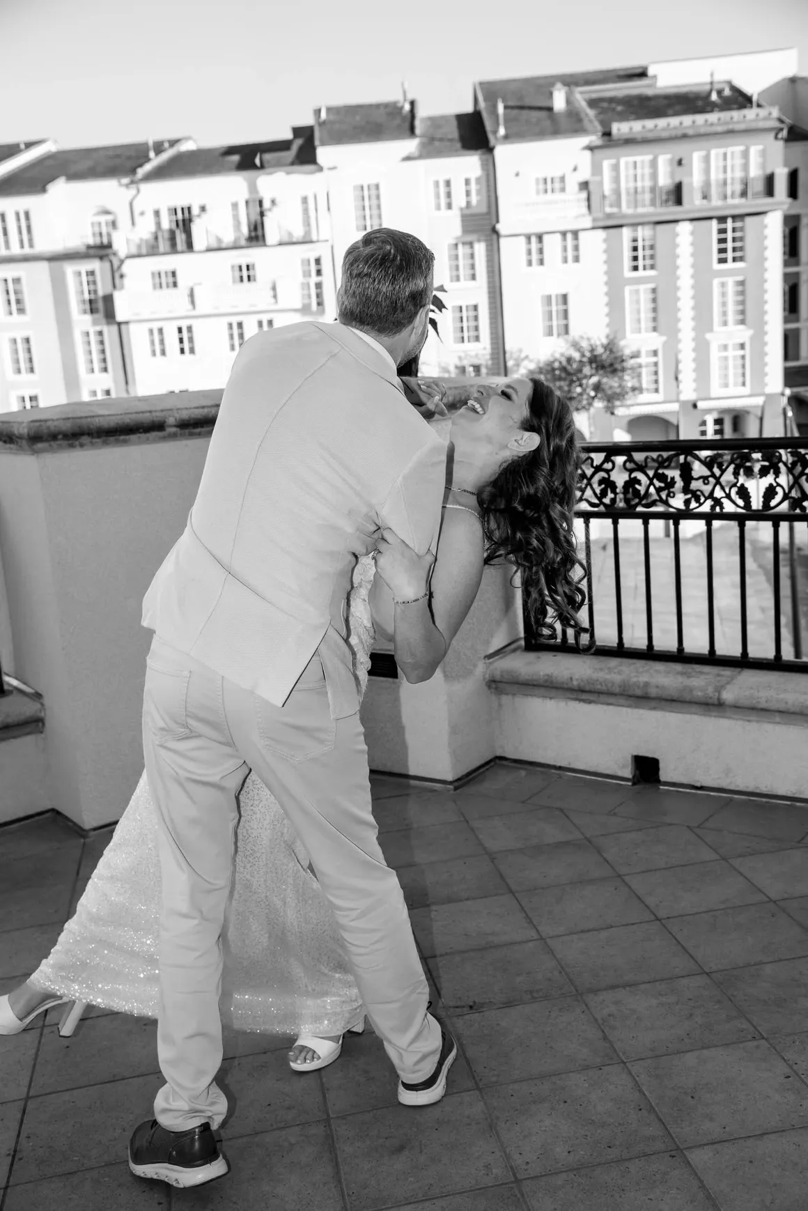 A person in a suit dips another person in a long dress on a balcony with a multi-story building in the background.
