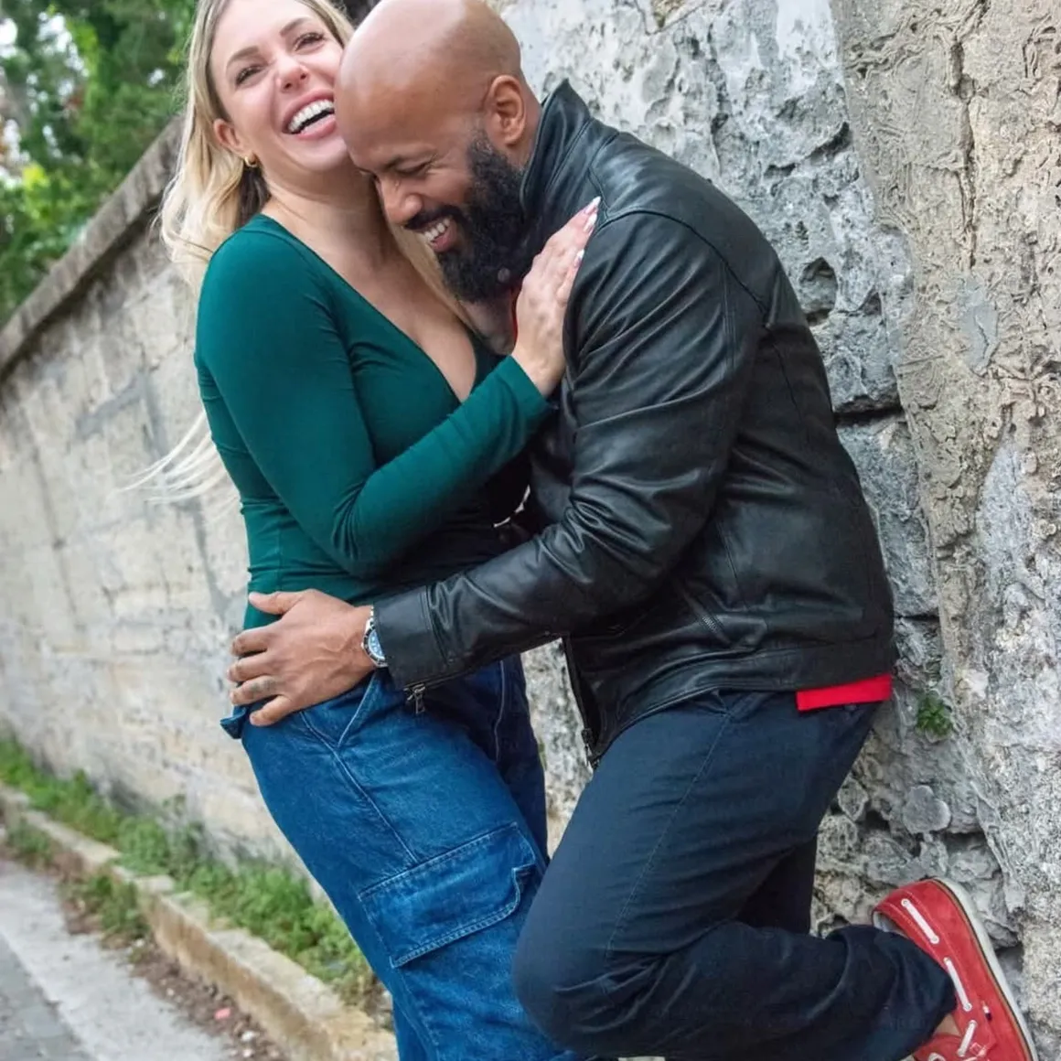 A smiling woman in a green top and blue jeans embraces a man in a black leather jacket near a stone wall.
