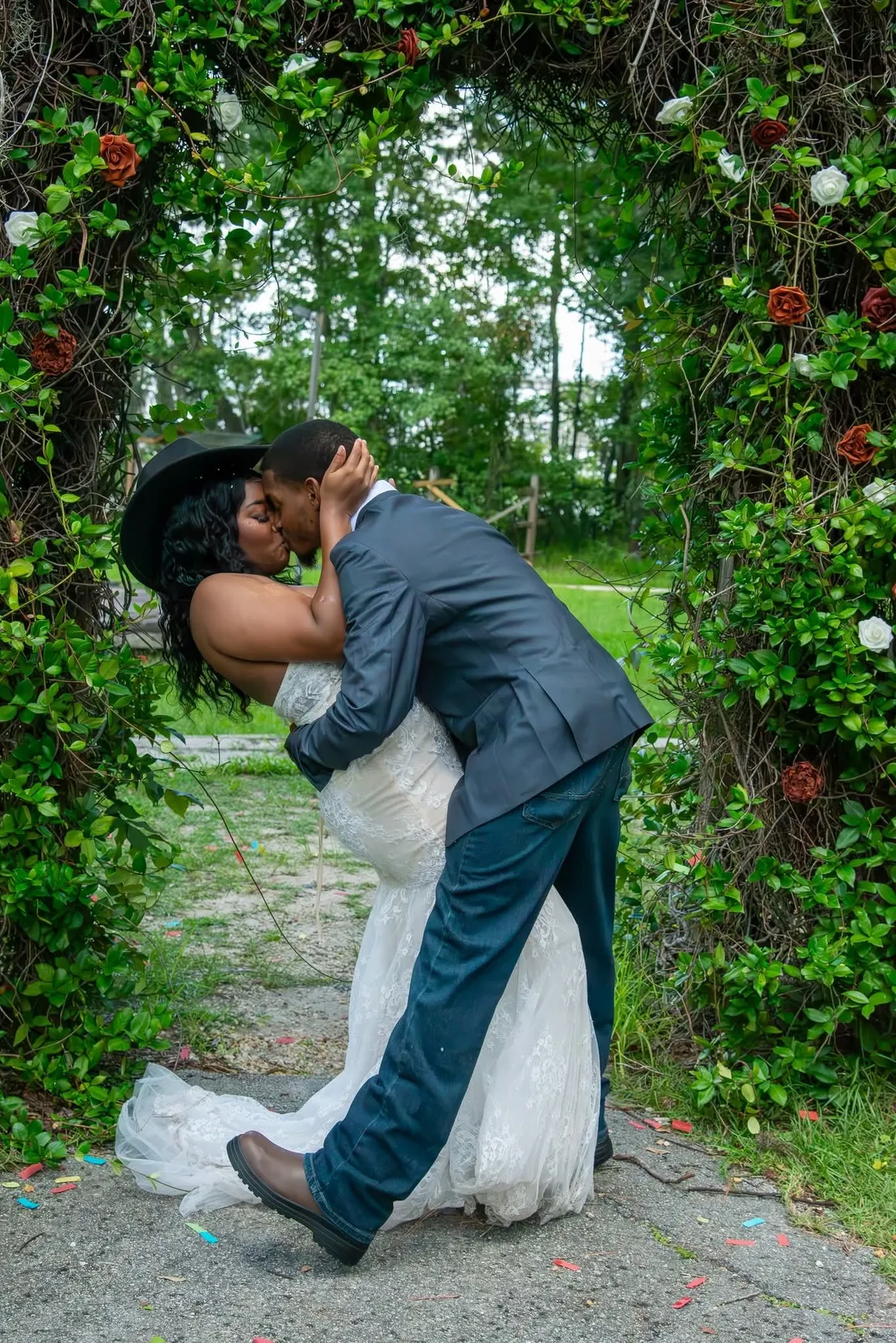 A couple kissing in a romantic dip beneath a leafy archway, surrounded by greenery and colorful flower accents.