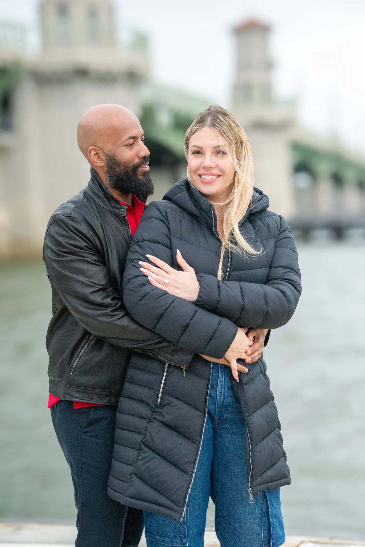 A couple embraces outdoors near a bridge, smiling toward the camera, wearing jackets and jeans.