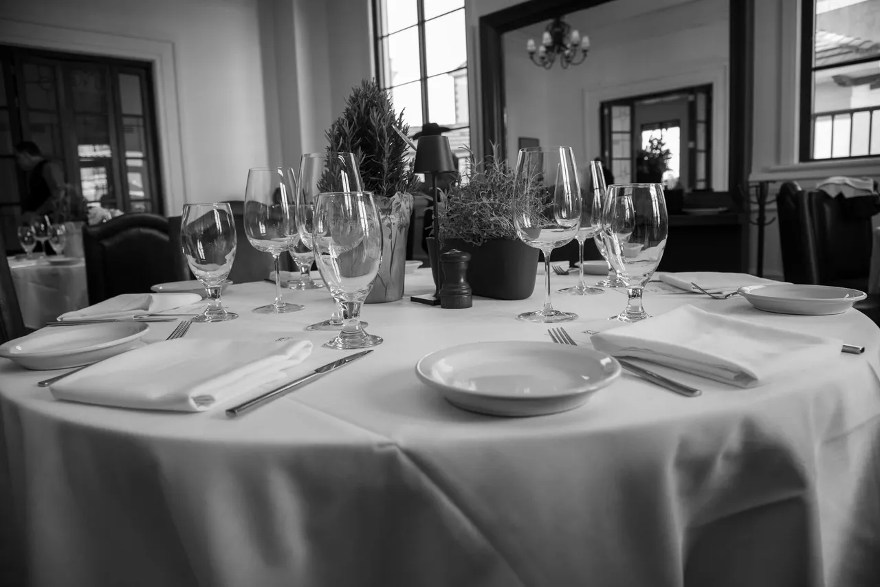 A formal dining table set for several people with white tablecloths, dinnerware, and wine glasses in a restaurant.
