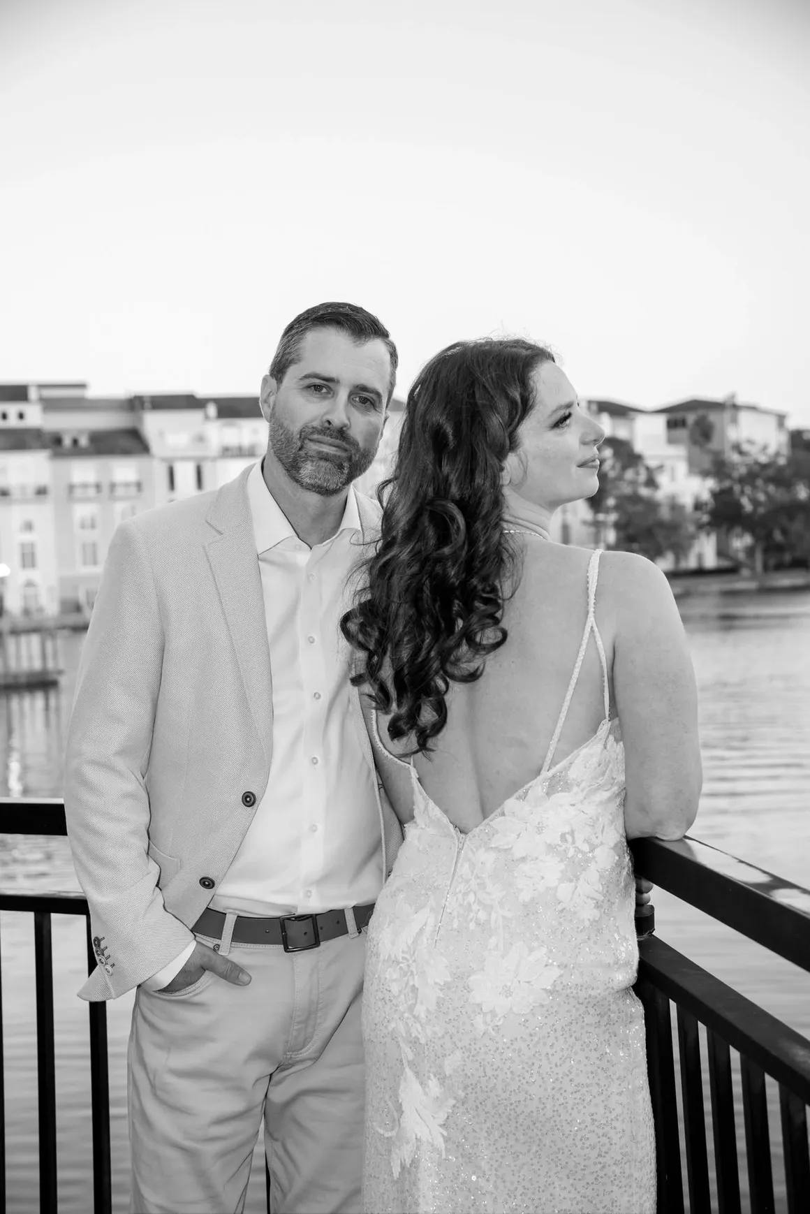 A couple in formal attire stands by a railing near a lake with buildings in the background, captured in monochrome.