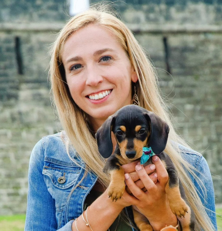 A blonde person smiles while holding a small black and tan dachshund puppy against a background of a stone wall.