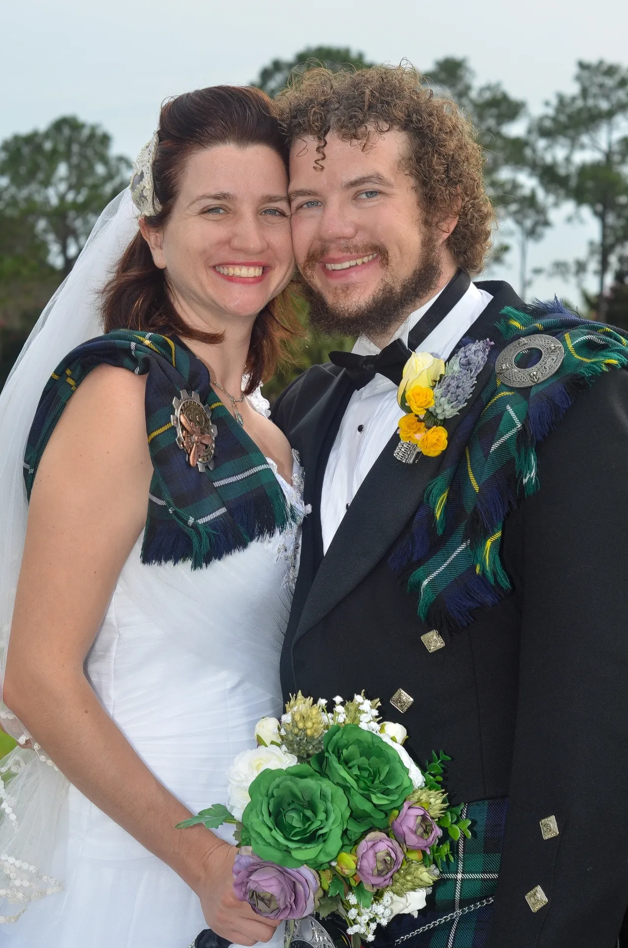 A couple smiles together wearing wedding attire with Scottish tartan sashes and a green floral bouquet.