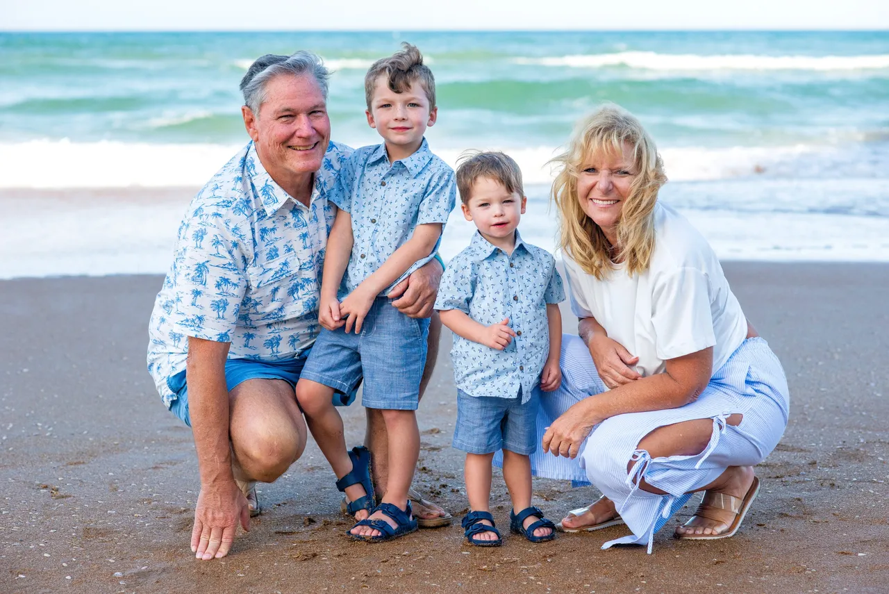 A family of four poses on a sandy beach by the ocean, wearing matching blue and white patterned outfits.