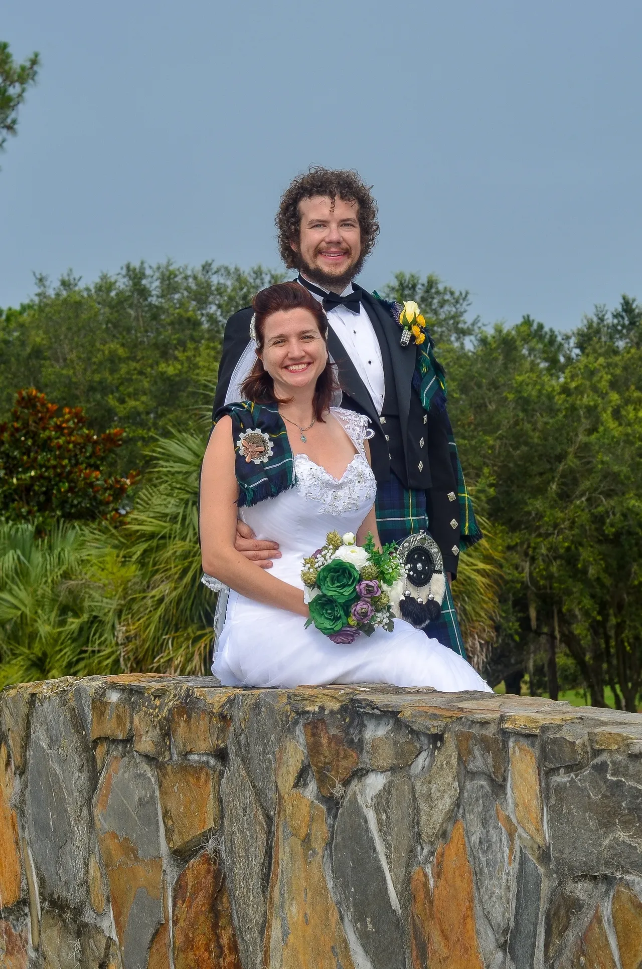 A bride in a white gown and a groom in a kilt and tuxedo pose for a portrait behind a stone wall in front of trees.