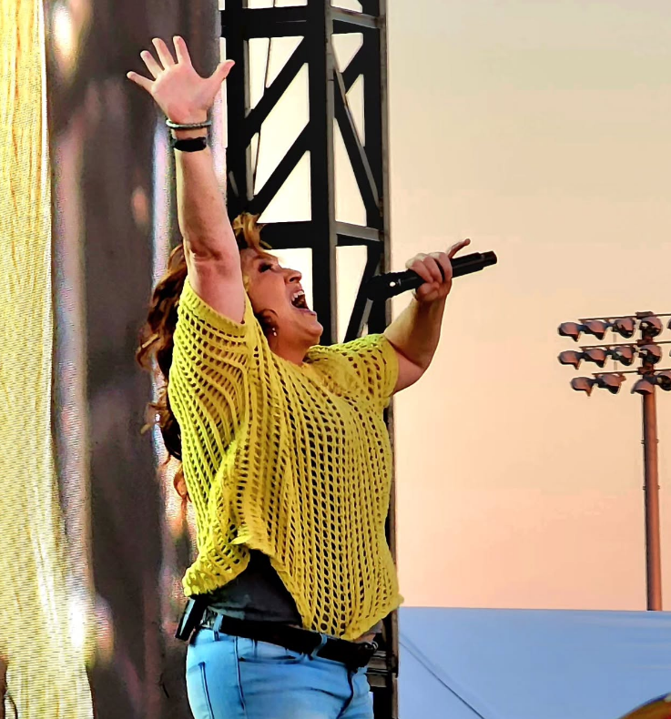 A person in a yellow crochet top and blue jeans performing on stage, arms raised with one hand holding a microphone.