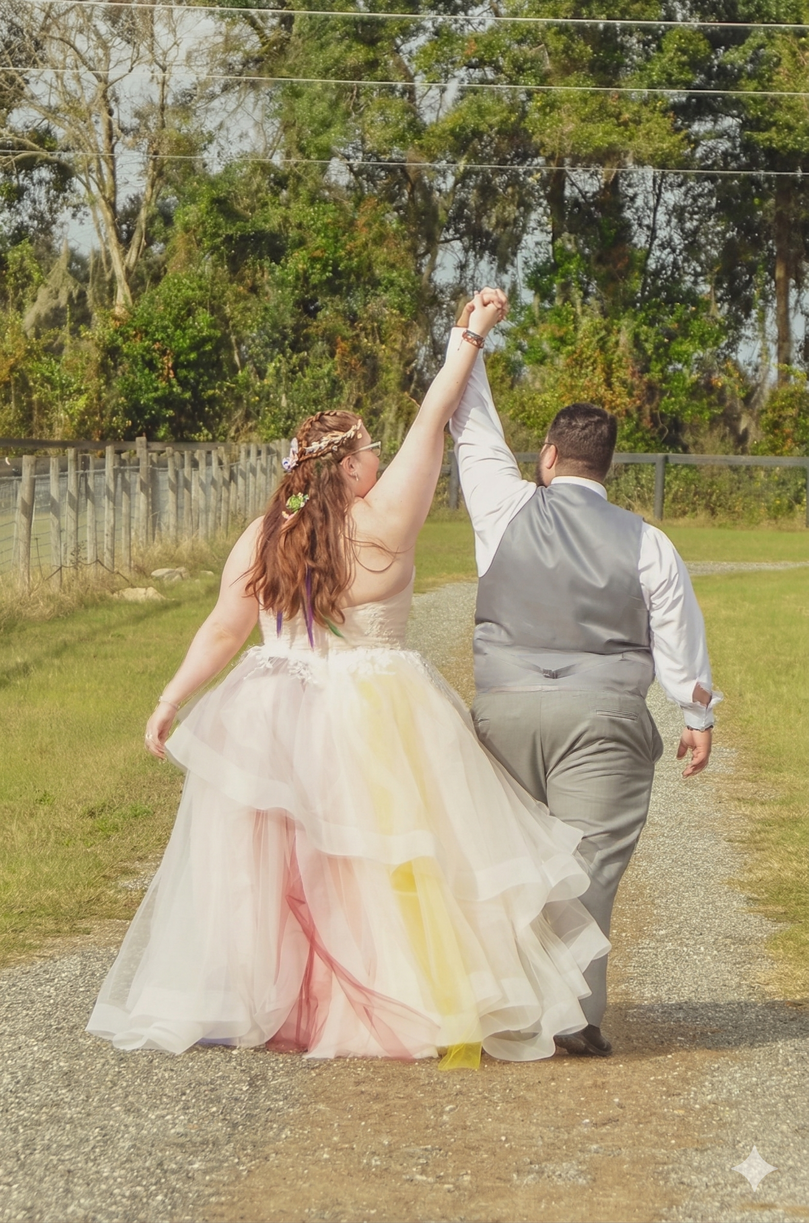 A person in a multi-colored tulle gown holds hands with a person in a grey vest, walking away on a path near a fence.