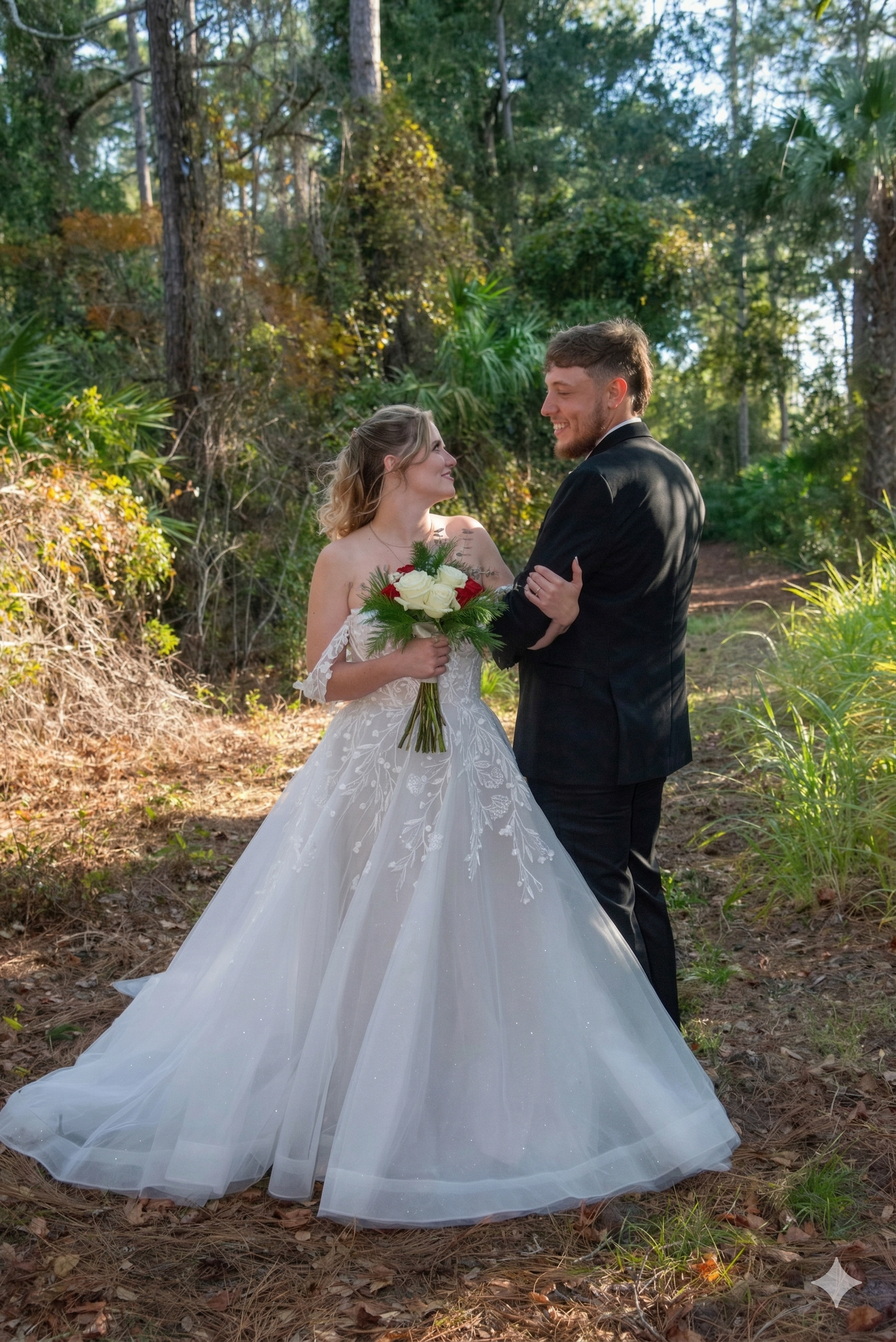 A bride in an off-the-shoulder gown holding a bouquet stands facing a groom in a black suit in a wooded area.