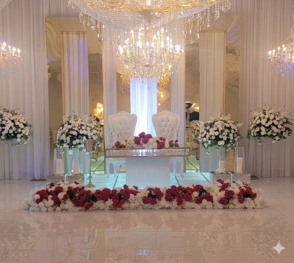 A white sweetheart table with two chairs, decorated with large floral arrangements and a base of red and white roses.