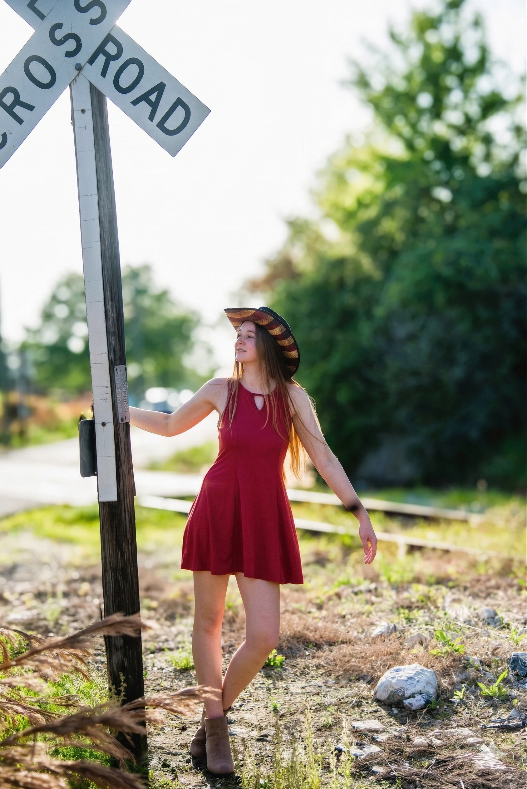 A person wearing a hat and red dress leaning against a railroad crossing sign on a gravel path near trees.