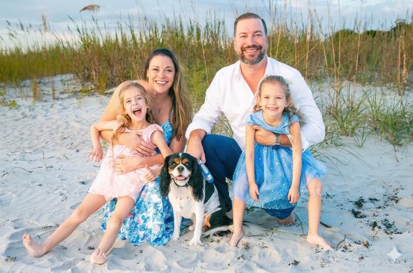 Family portrait on a sandy beach at sunset, including a dog sitting with them amidst tall beach grass.