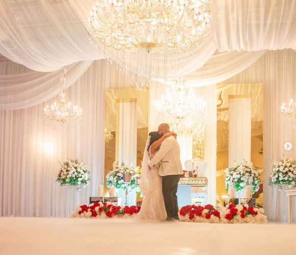 A couple embraces during their first dance in a formal venue decorated with white drapery, chandeliers, and red roses.