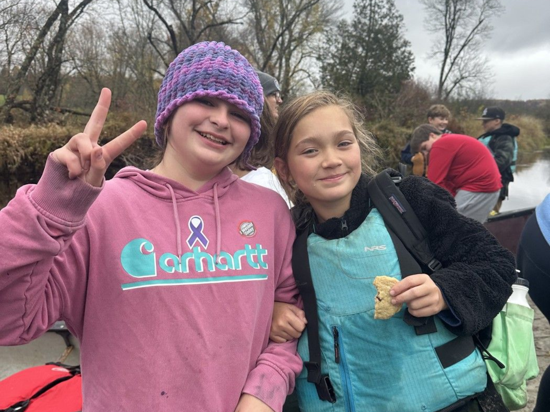 Two smiling kids outdoors; one in a pink Carhartt hoodie and purple hat flashes a peace sign.