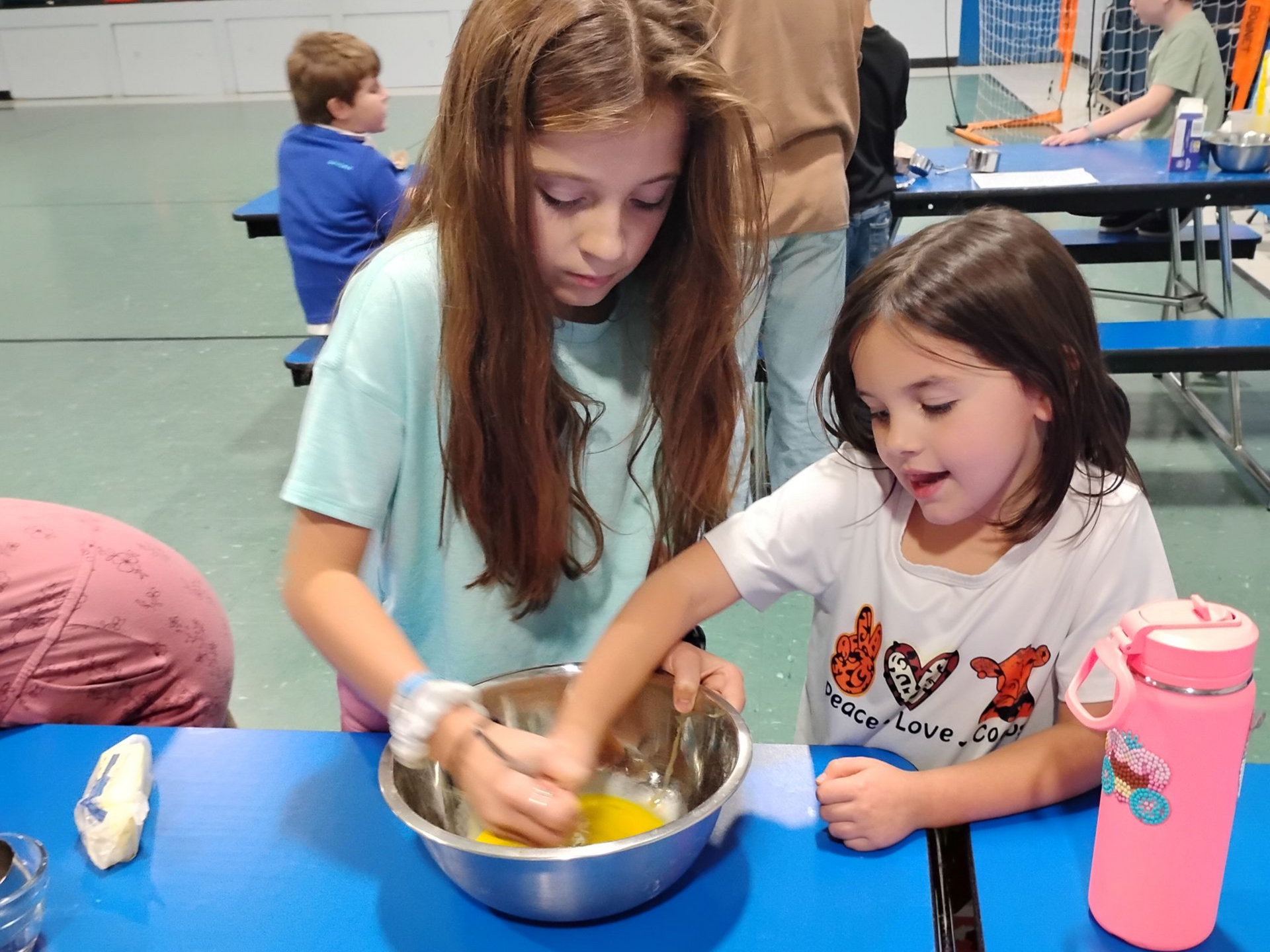 Two children mixing ingredients in a metal bowl at a blue table during a classroom activity