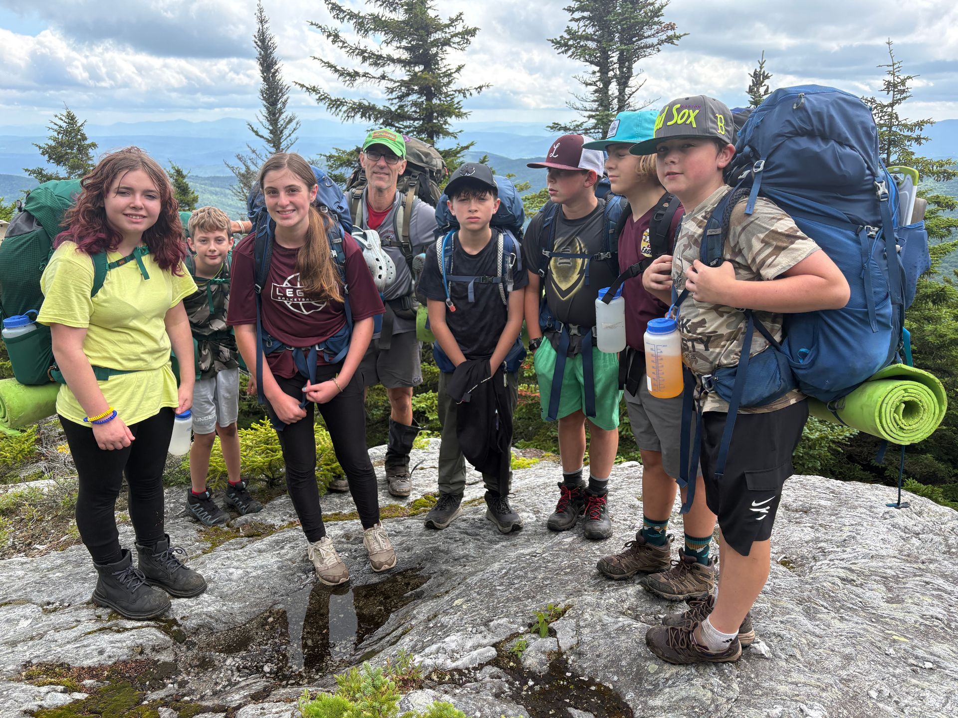 Six hikers in colorful backpacks pose on a rocky mountain trail with trees and cloudy sky.