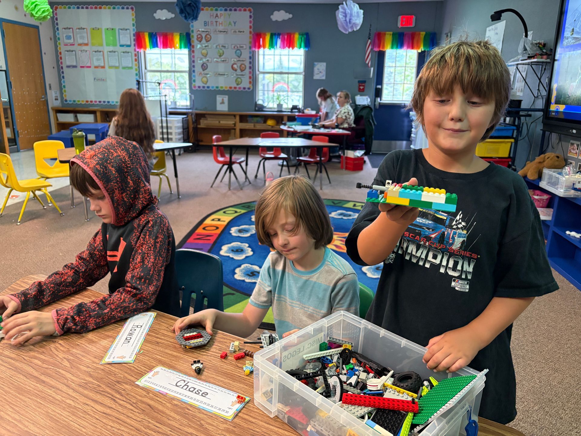 Children playing with toys at a classroom table, with colorful walls and chairs in the background