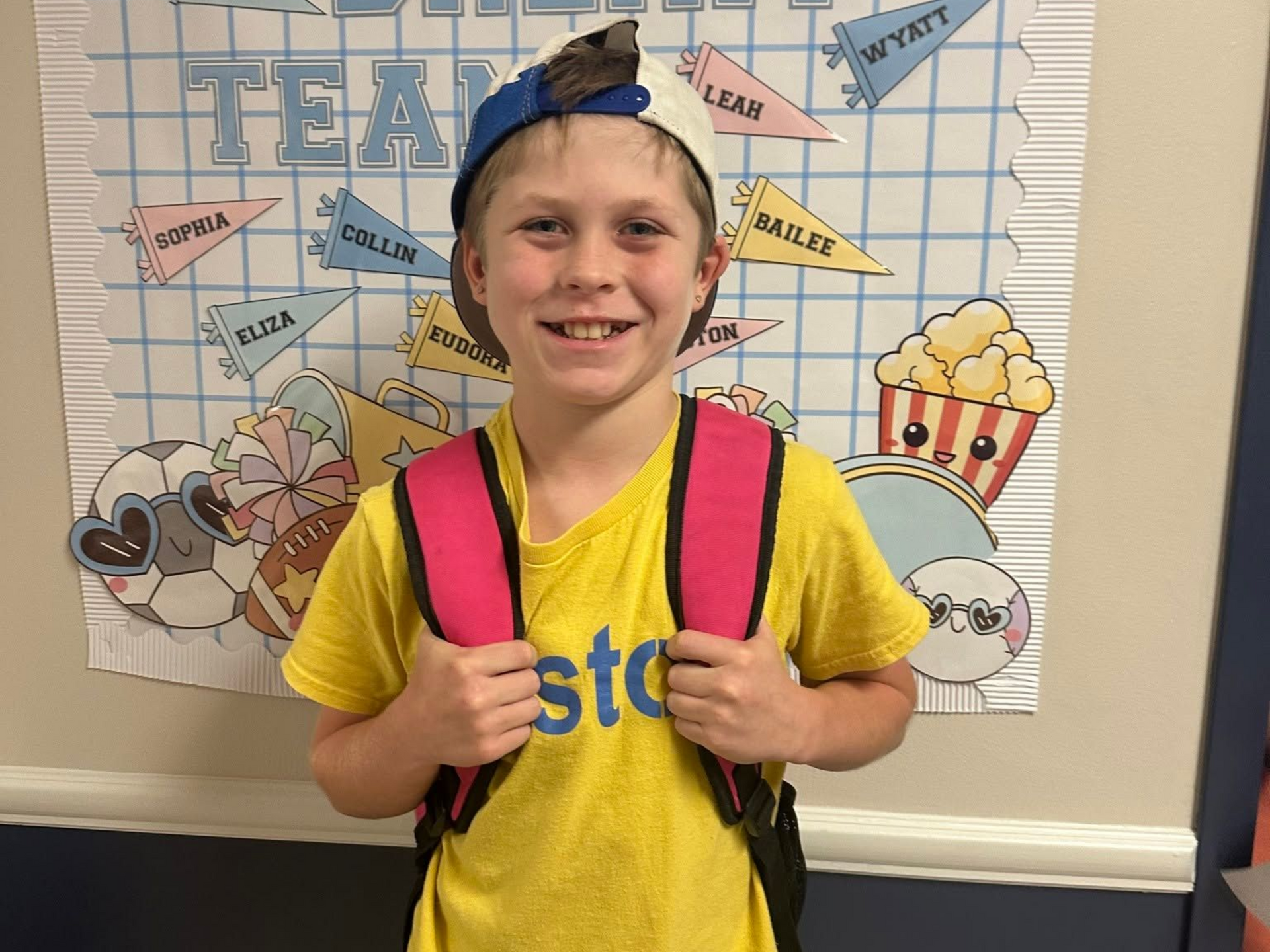 Smiling child with backpack and cap standing in front of a classroom welcome board