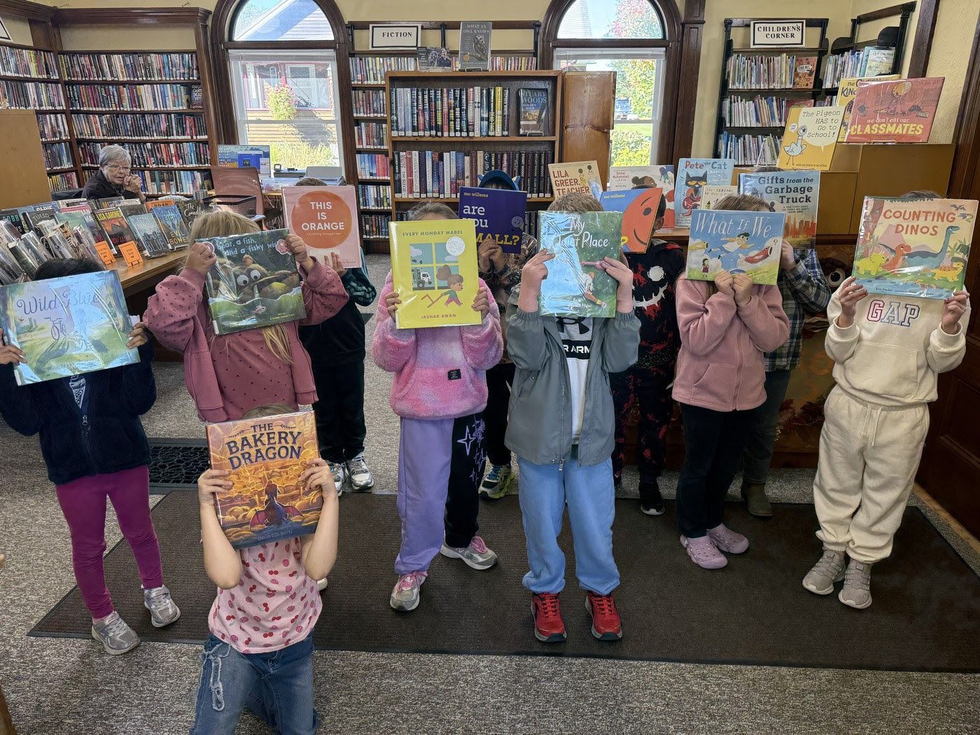 Children holding up colorful books in a library, standing in a row with bookshelves behind them