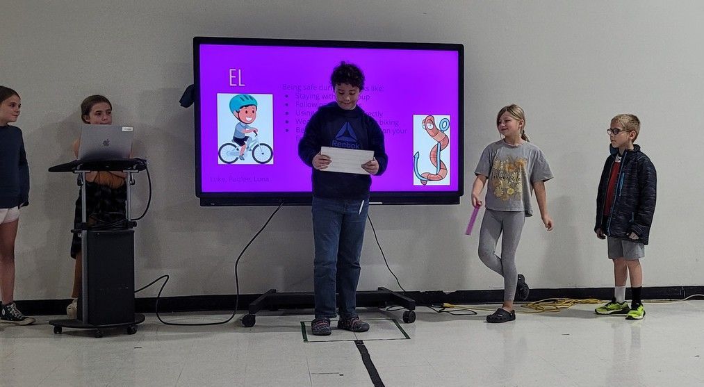 Children stand on a stage in front of a large purple screen during a presentation.
