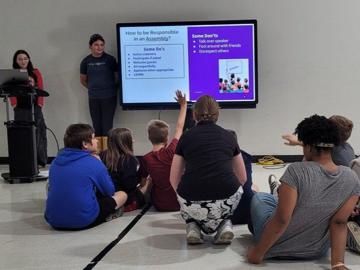 Audience seated in a room, listening to a presentation on twin wall screens