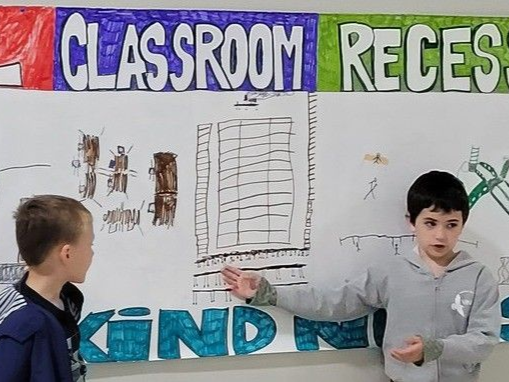 Classroom wall display with posters; two children stand in front of it, one pointing at the board.