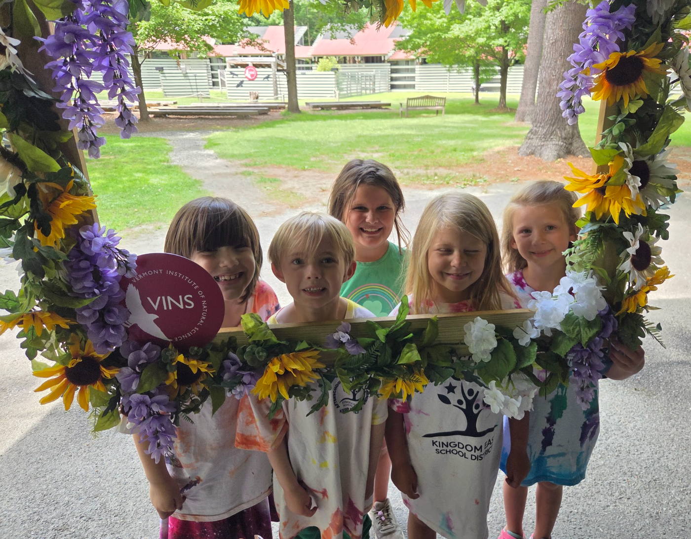 Four children smiling behind a purple and yellow flower wreath frame outdoors, holding a purple balloon