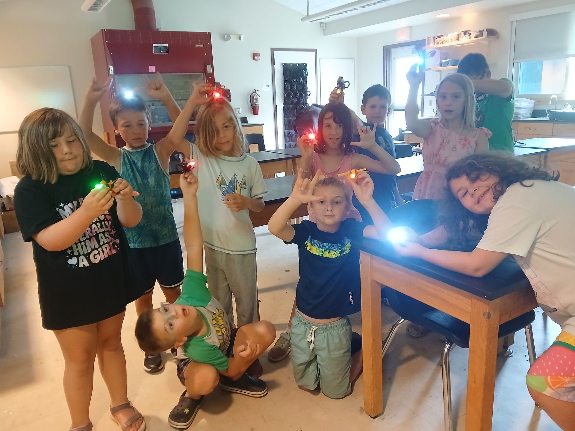 Children holding flashlights pose around a table in a bright room, with one girl lying across the table.